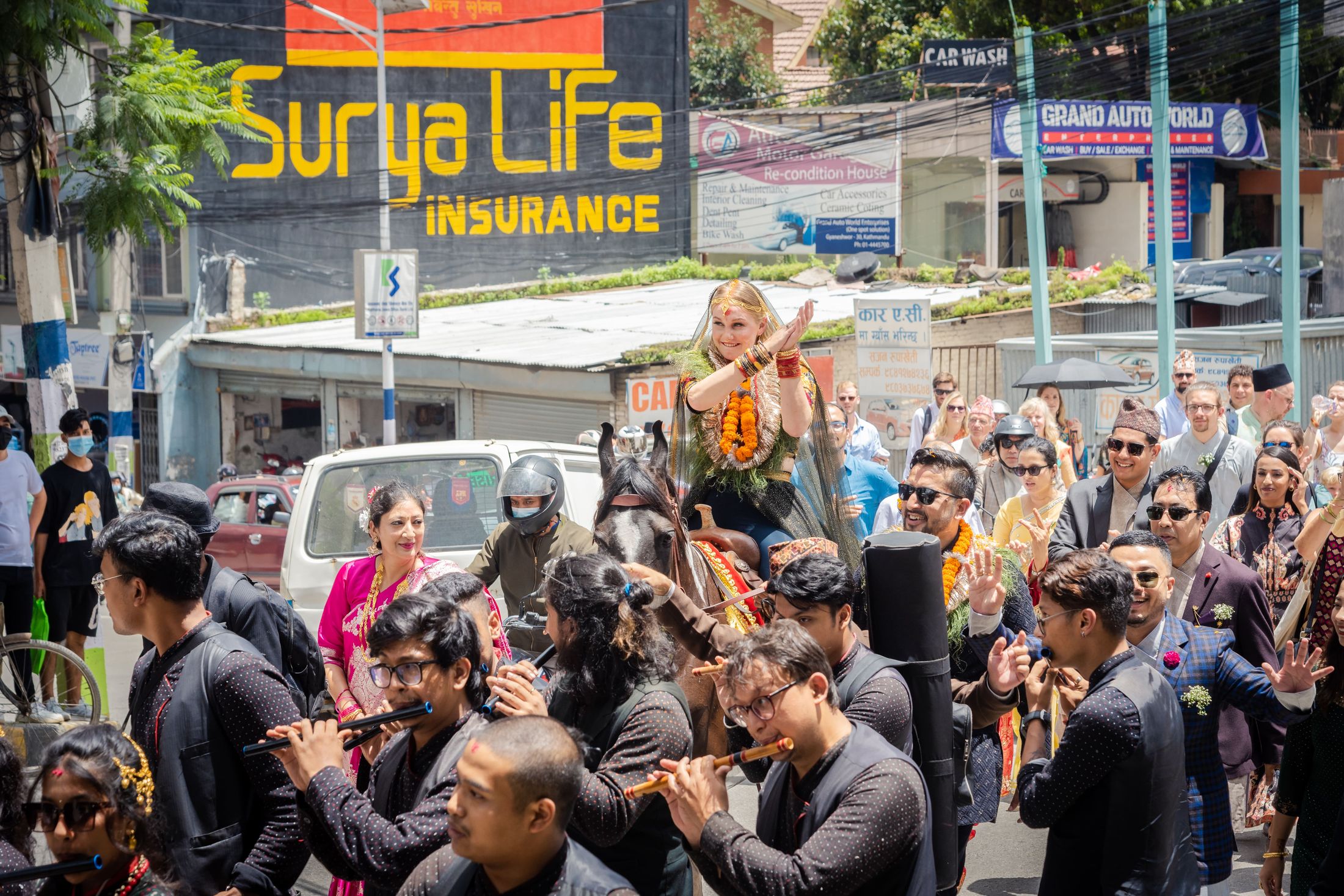 Street baraat procession for Jana and Raymon with a live band playing traditional instruments, a Surya Life Insurance signboard visible behind the crowd