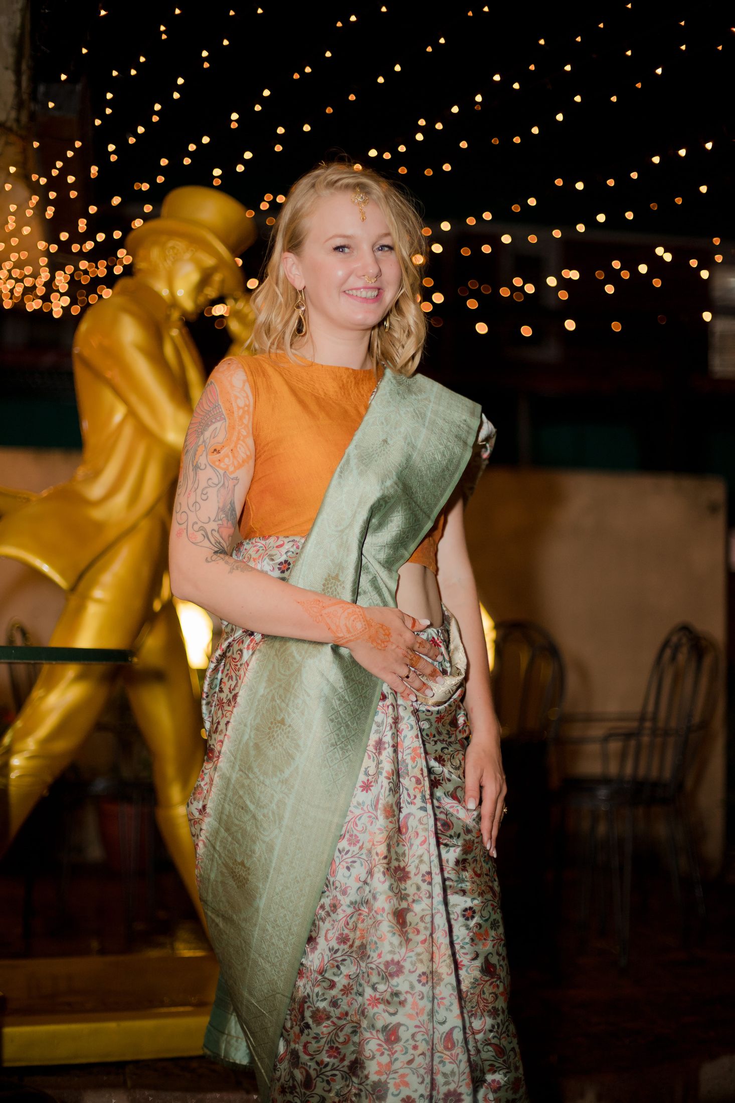 Jana standing alone near a golden statue at night with string lights overhead