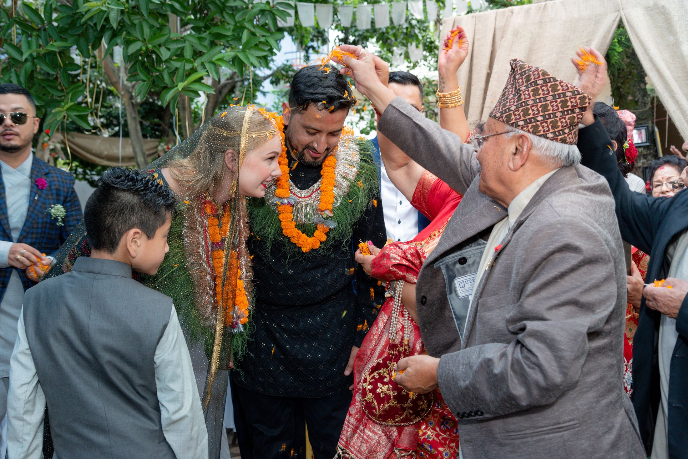 Jana and Raymon walking together under a tan fabric canopy, wearing marigold garlands