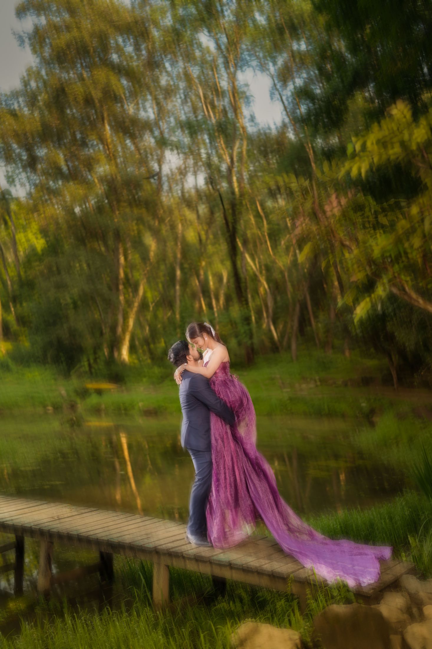 Bride in a flowing purple gown embracing groom on a wooden footbridge surrounded by greenery