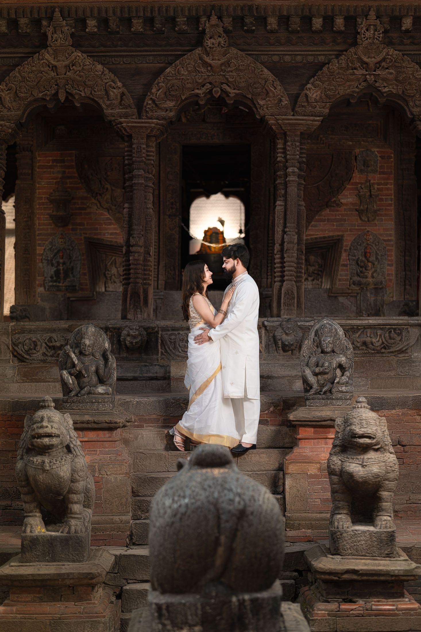Bride and groom standing in an ornate carved temple doorway flanked by stone lion statues at dusk