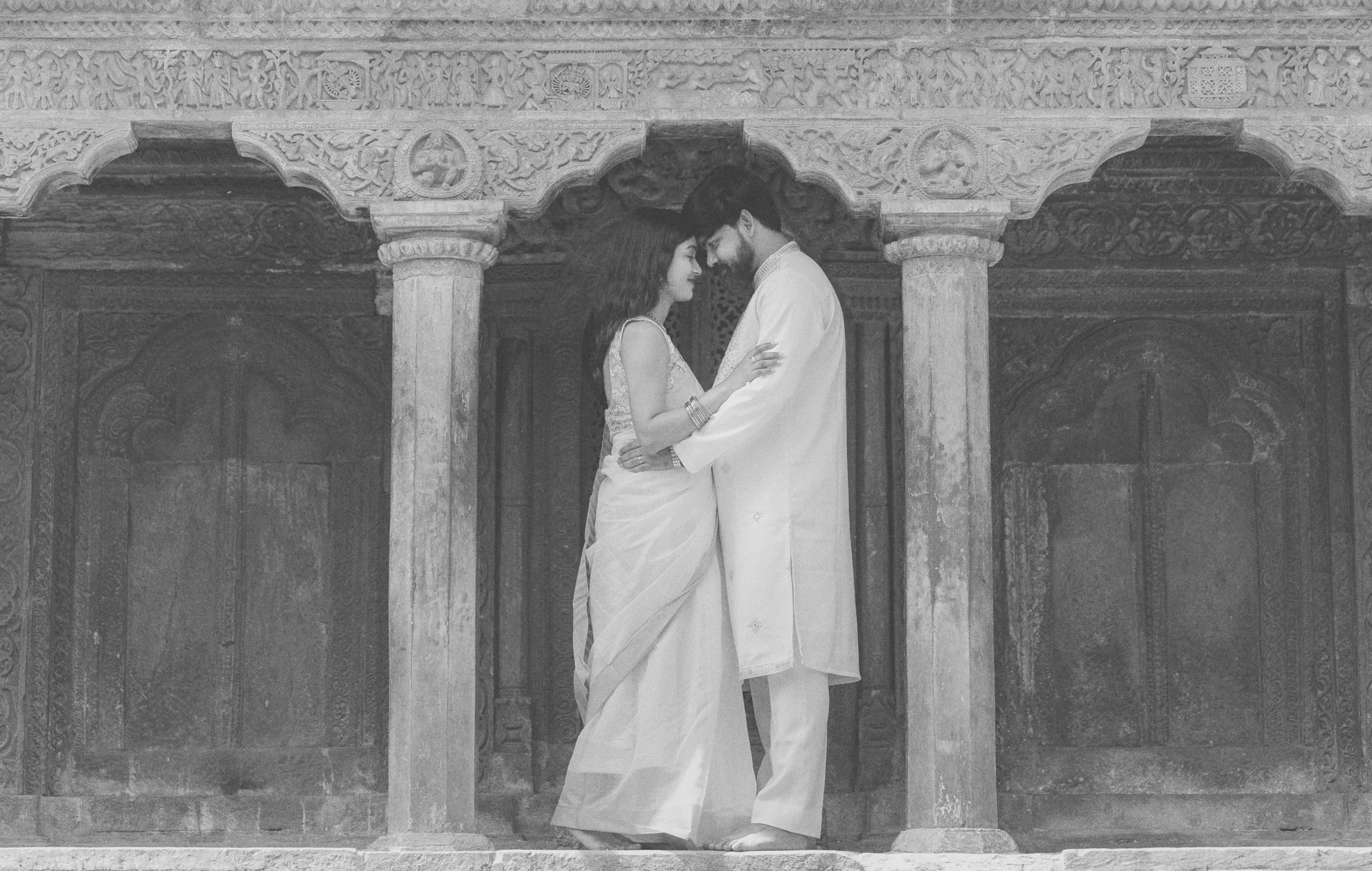 Black and white photo of bride and groom embracing beneath ornate temple stone pillars and arches