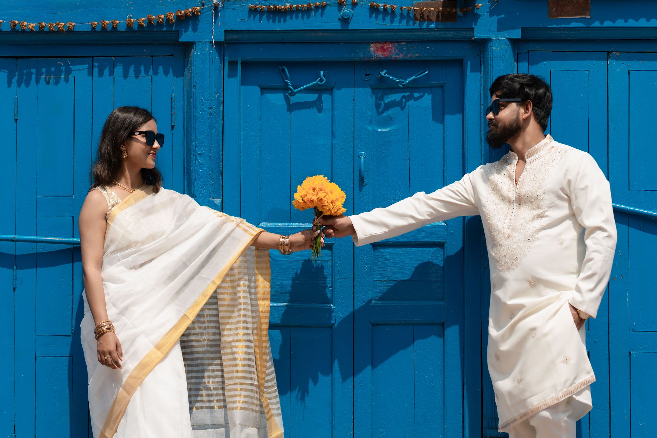 Bride and groom in cream traditional outfits holding a marigold garland in front of a blue wooden door