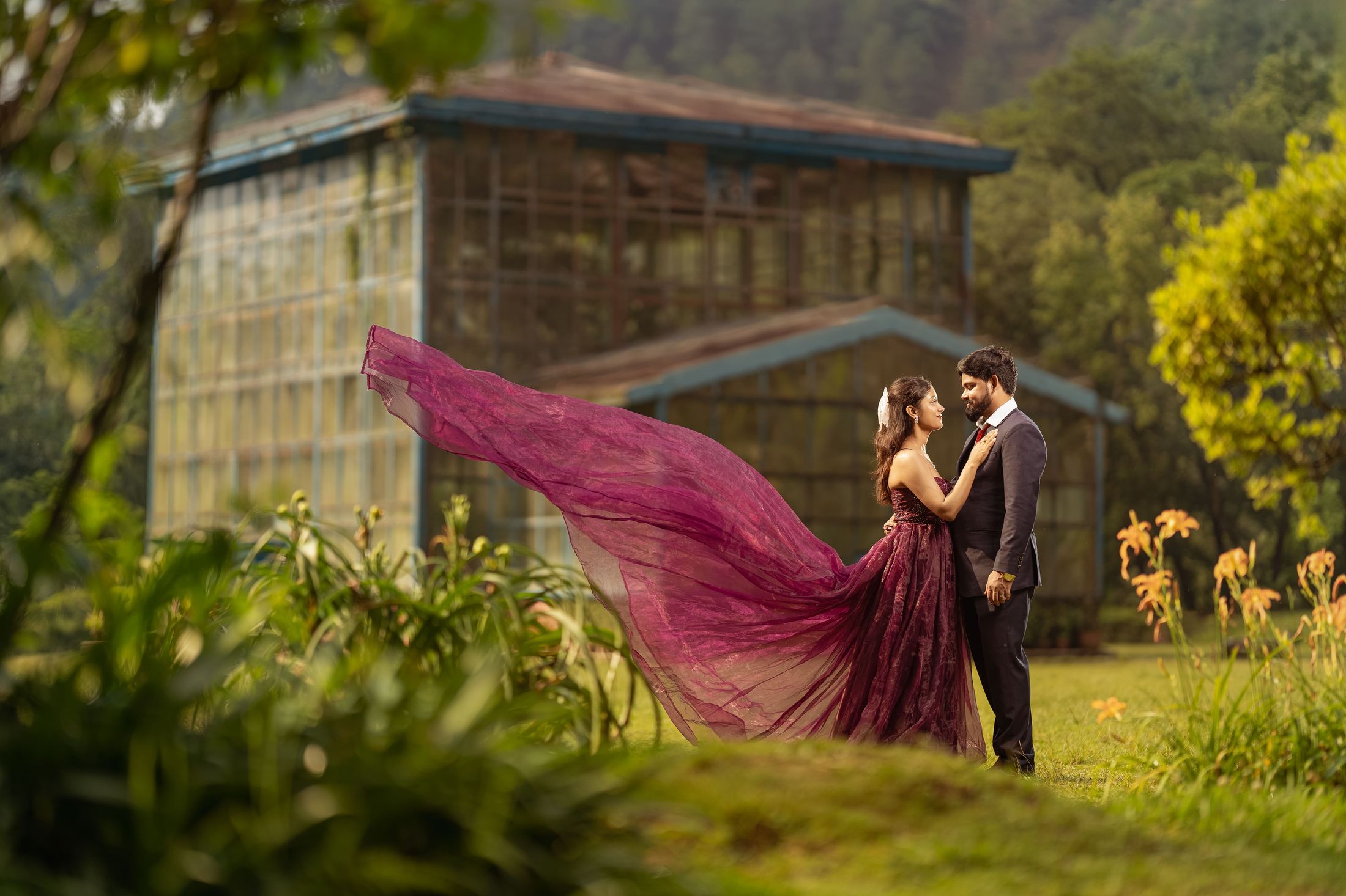 Bride in a flowing wine-colored gown and groom standing together in a garden greenhouse setting