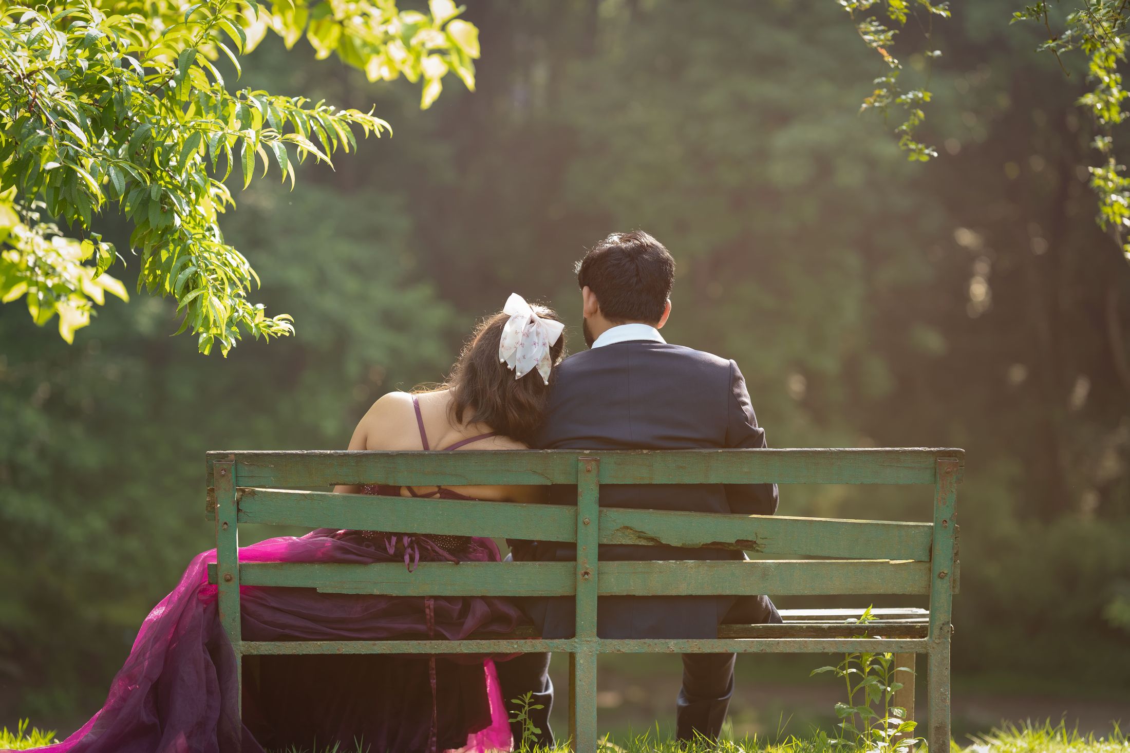 Bride and groom sitting together on a park bench viewed from behind, her gown draped across the ground
