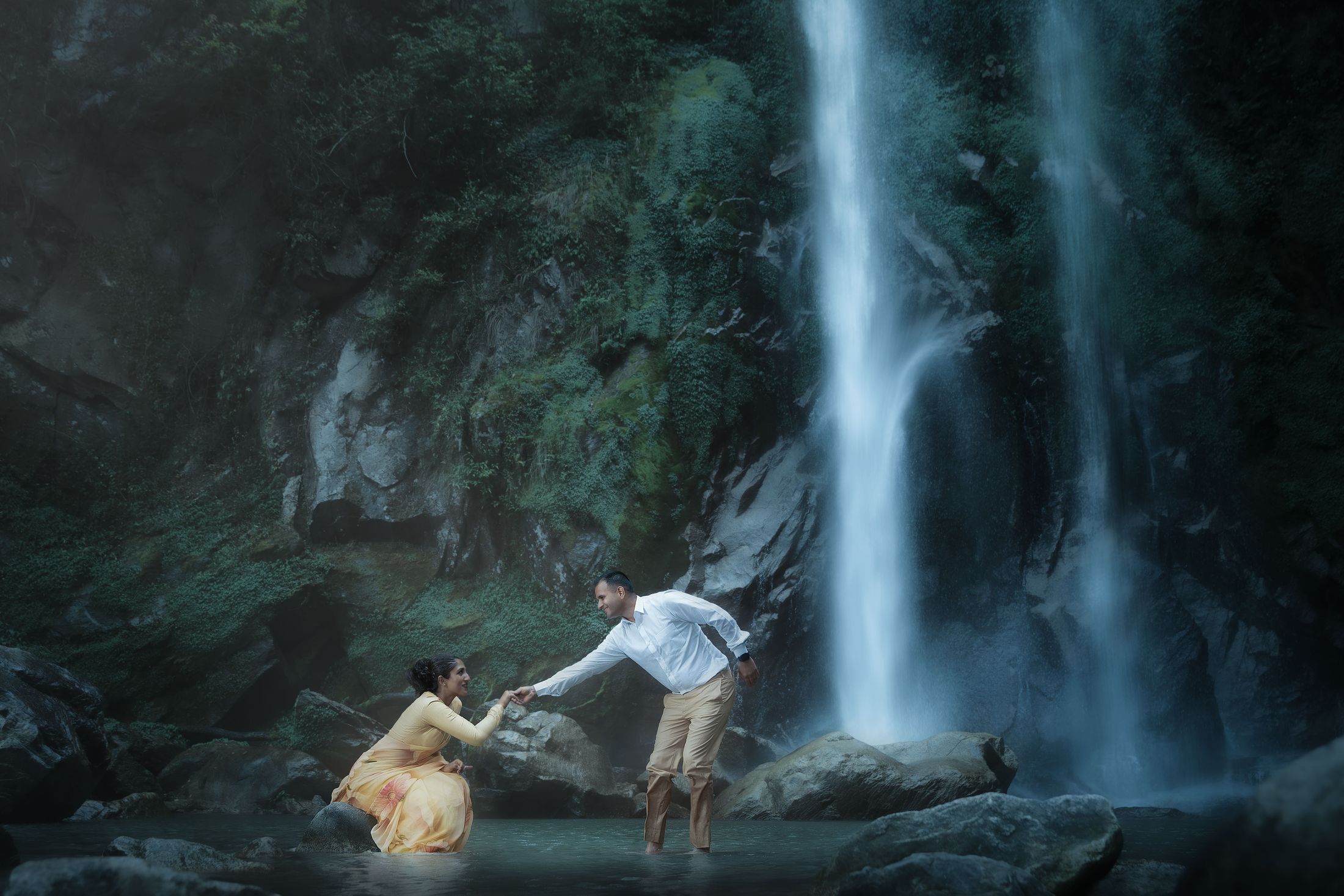 Krishna helping Disuja climb down rocks in front of a large waterfall during their pre-wedding shoot, she in a yellow dress