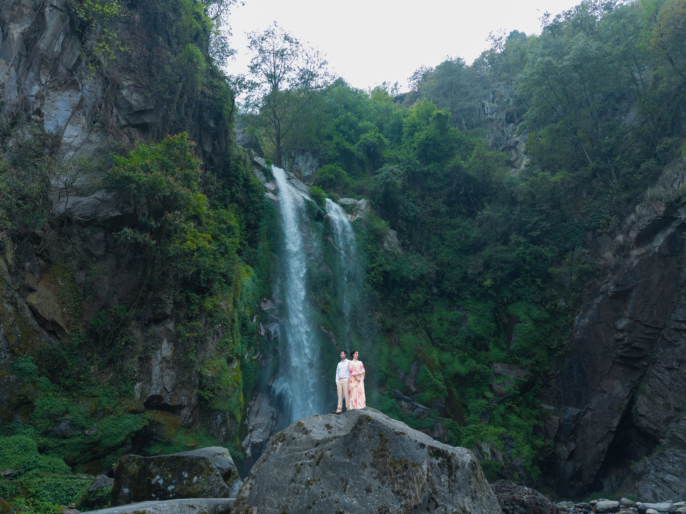 Wide shot of Disuja and Krishna standing on a rock ledge dwarfed by a tall waterfall surrounded by green cliffs