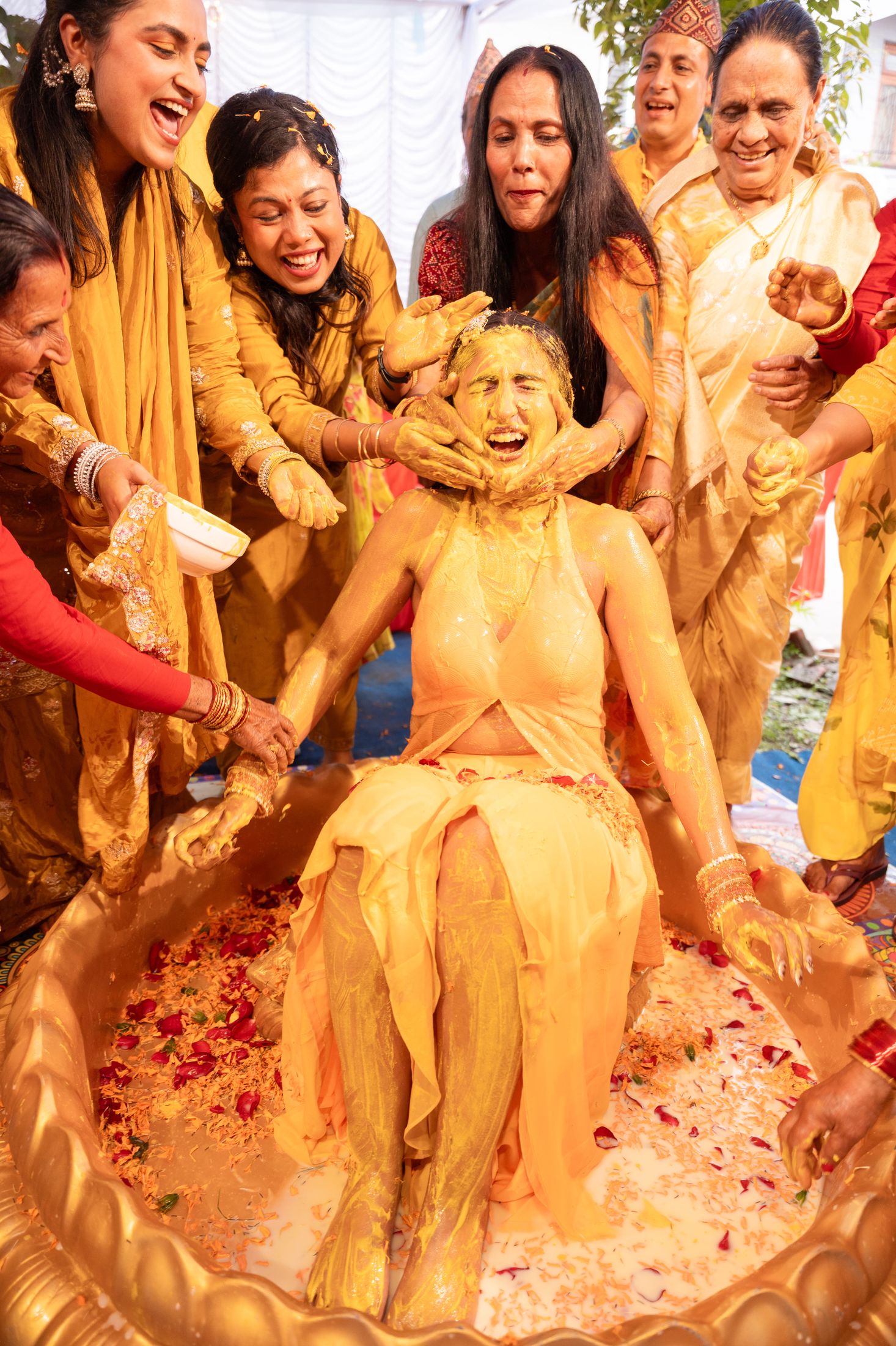 Disuja seated in a golden basin being covered in turmeric paste by female family members during her haldi ceremony