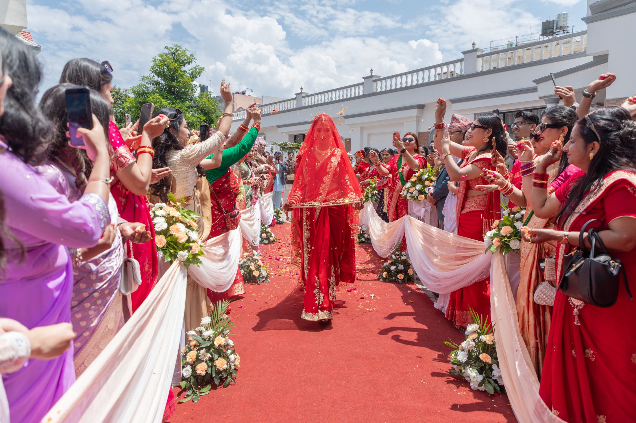Disuja walking down a red carpet aisle under a red veil held up by women, guests celebrating on either side