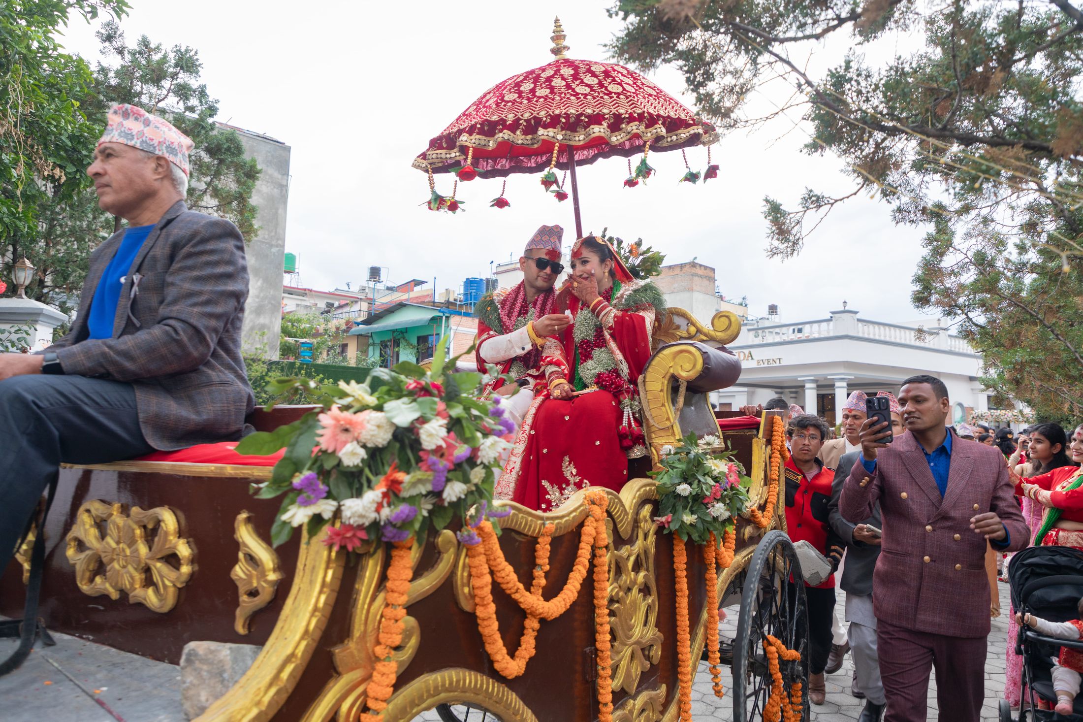 Couple riding in an ornate flower-decorated carriage with a red umbrella canopy through the street