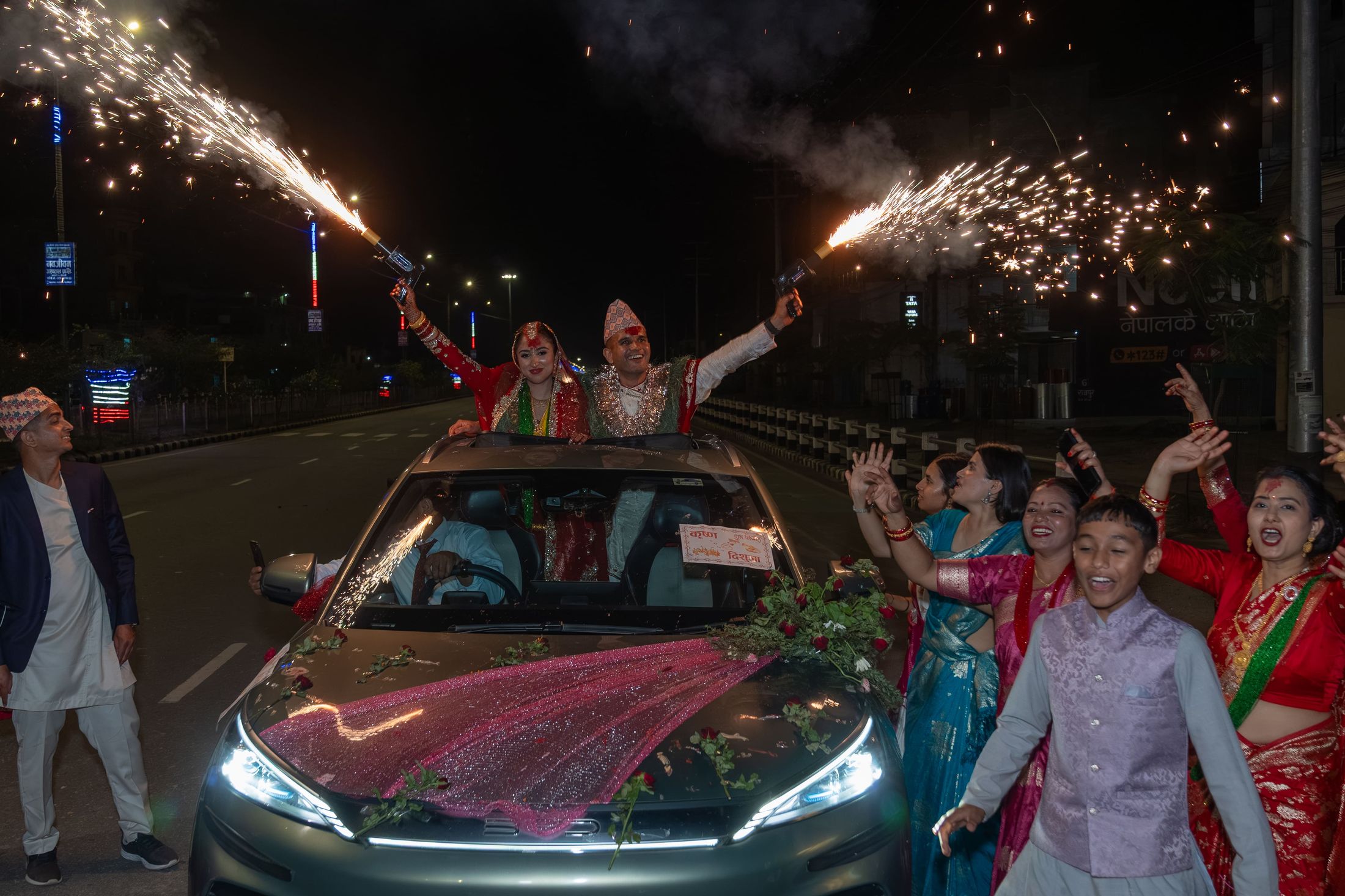 Couple celebrating at night through a car sunroof surrounded by sparklers and family