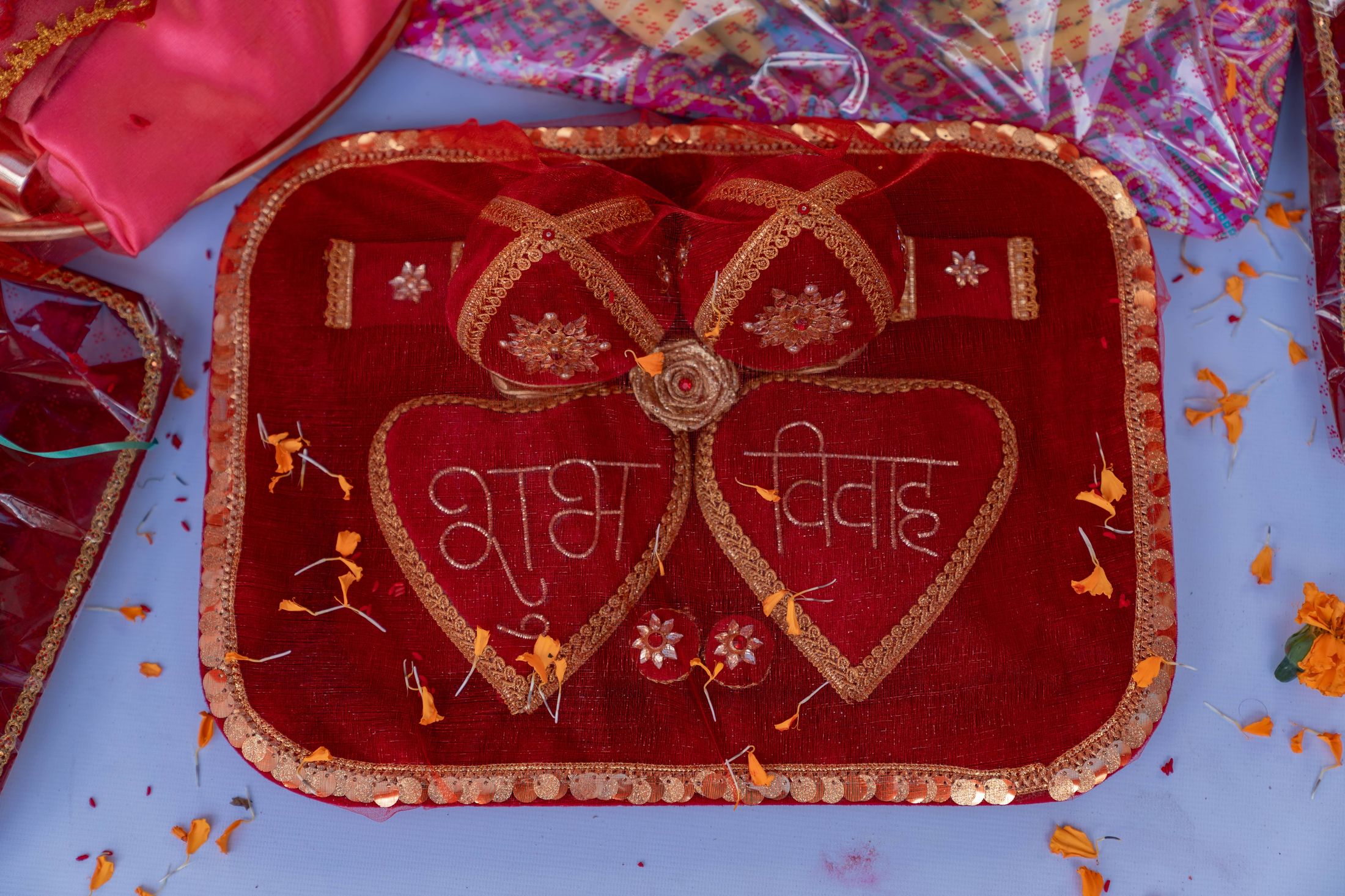 Red heart-shaped ceremonial invitation card embroidered with Shubh Vivah text, surrounded by scattered flower petals