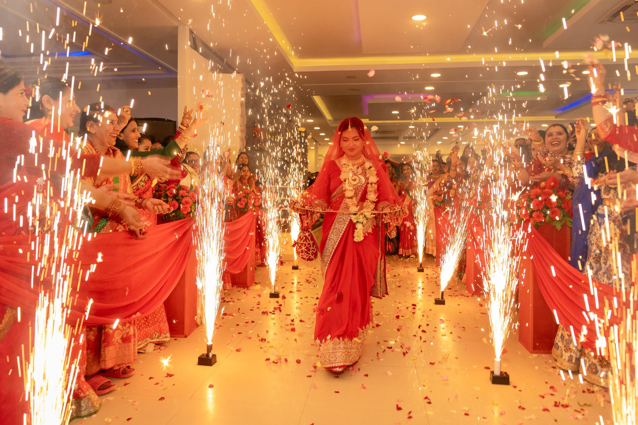 Inusha walking down the aisle in a red saree with a floral garland, flanked by cold spark fountains as family celebrates