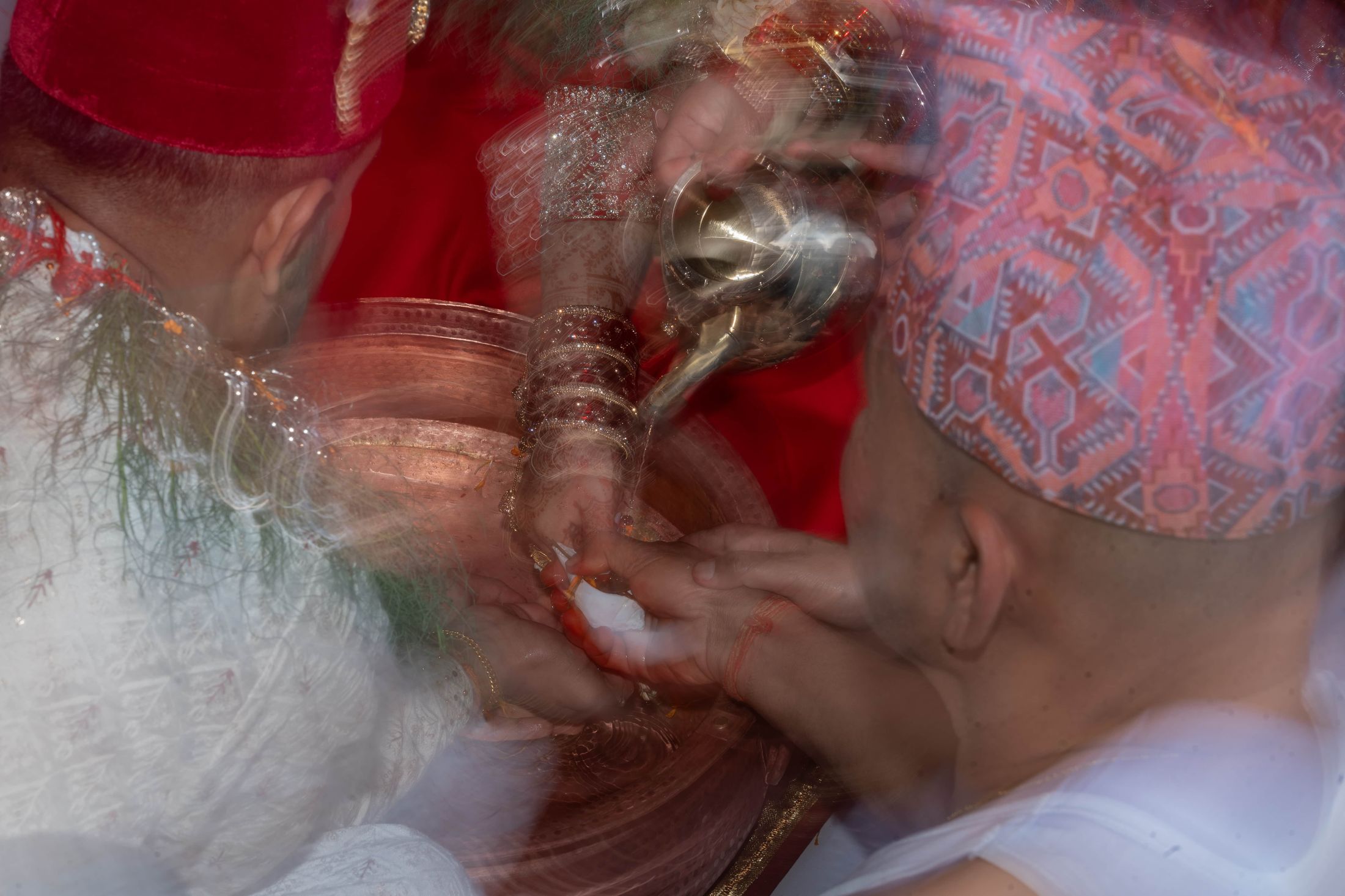 Motion-blurred shot of Prashid pouring rice into Inusha's hands during a ceremony, both wearing traditional Nepali topi
