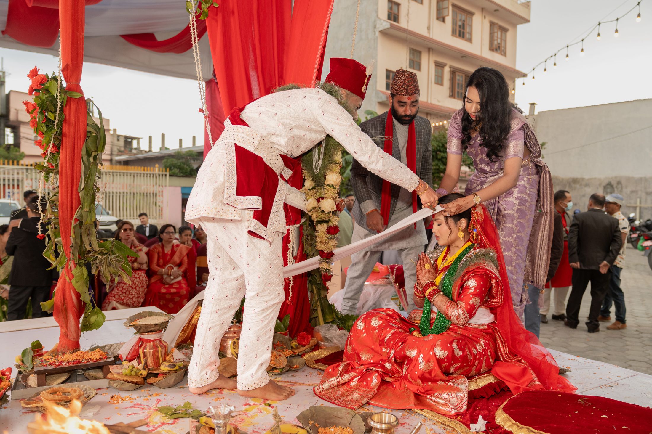 Prashid placing a ceremonial item on Inusha's forehead as she sits under a red and white canopy with an officiant beside them