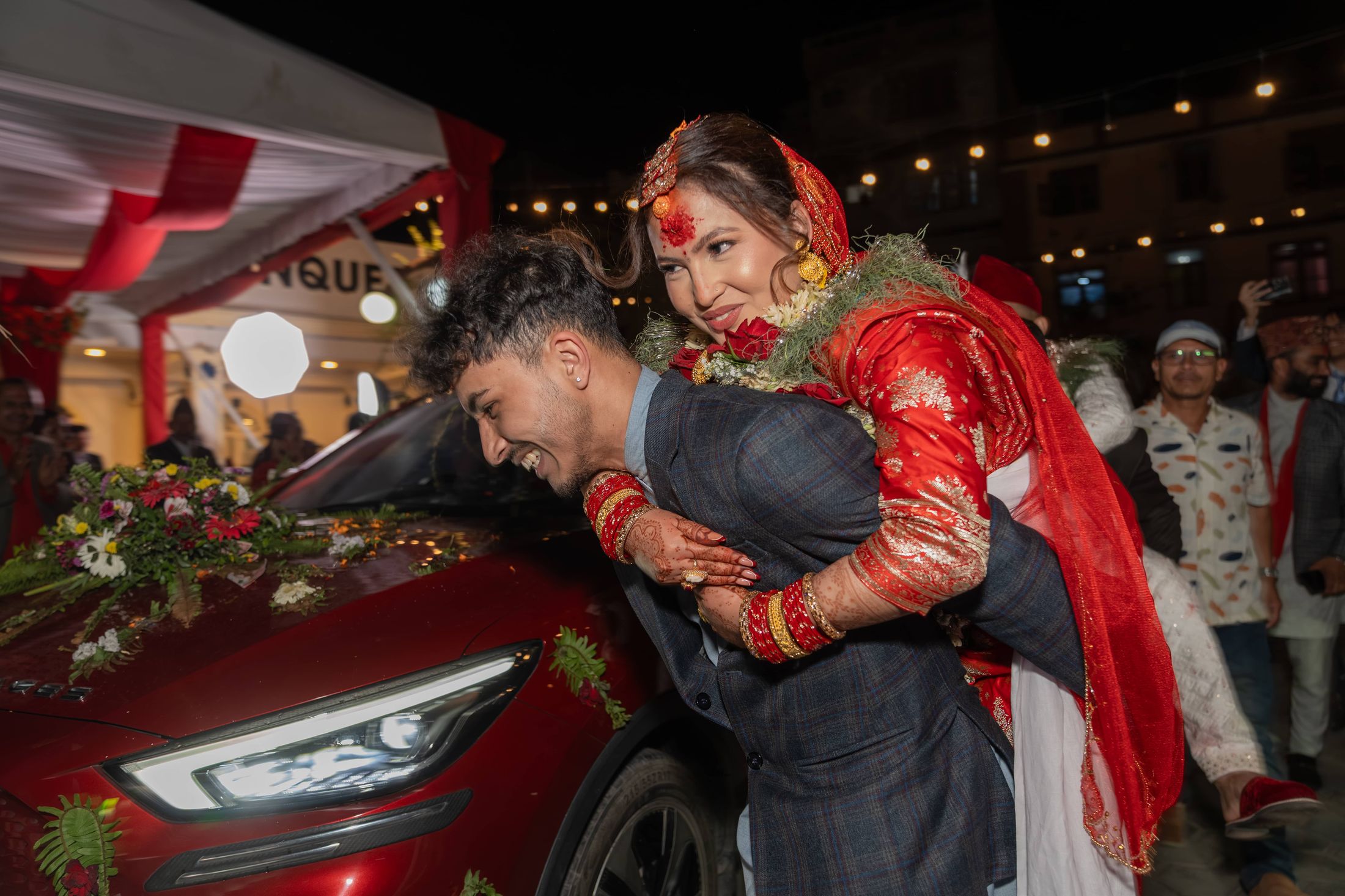 Prashid playfully carrying Inusha piggyback beside a red car at night as guests laugh around them