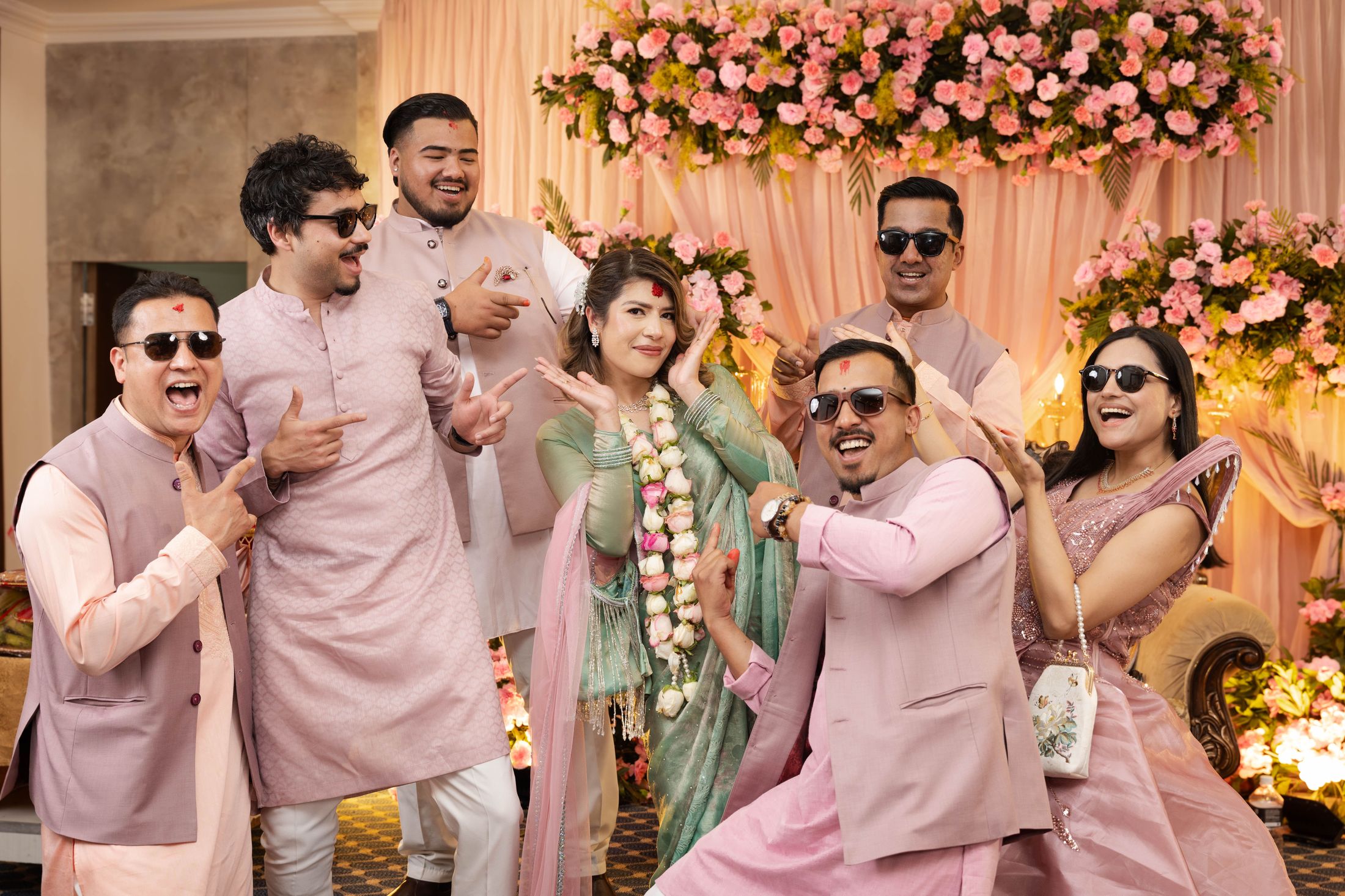 Group of five friends posing playfully with sunglasses and hand gestures around Suprabha against a pink floral backdrop