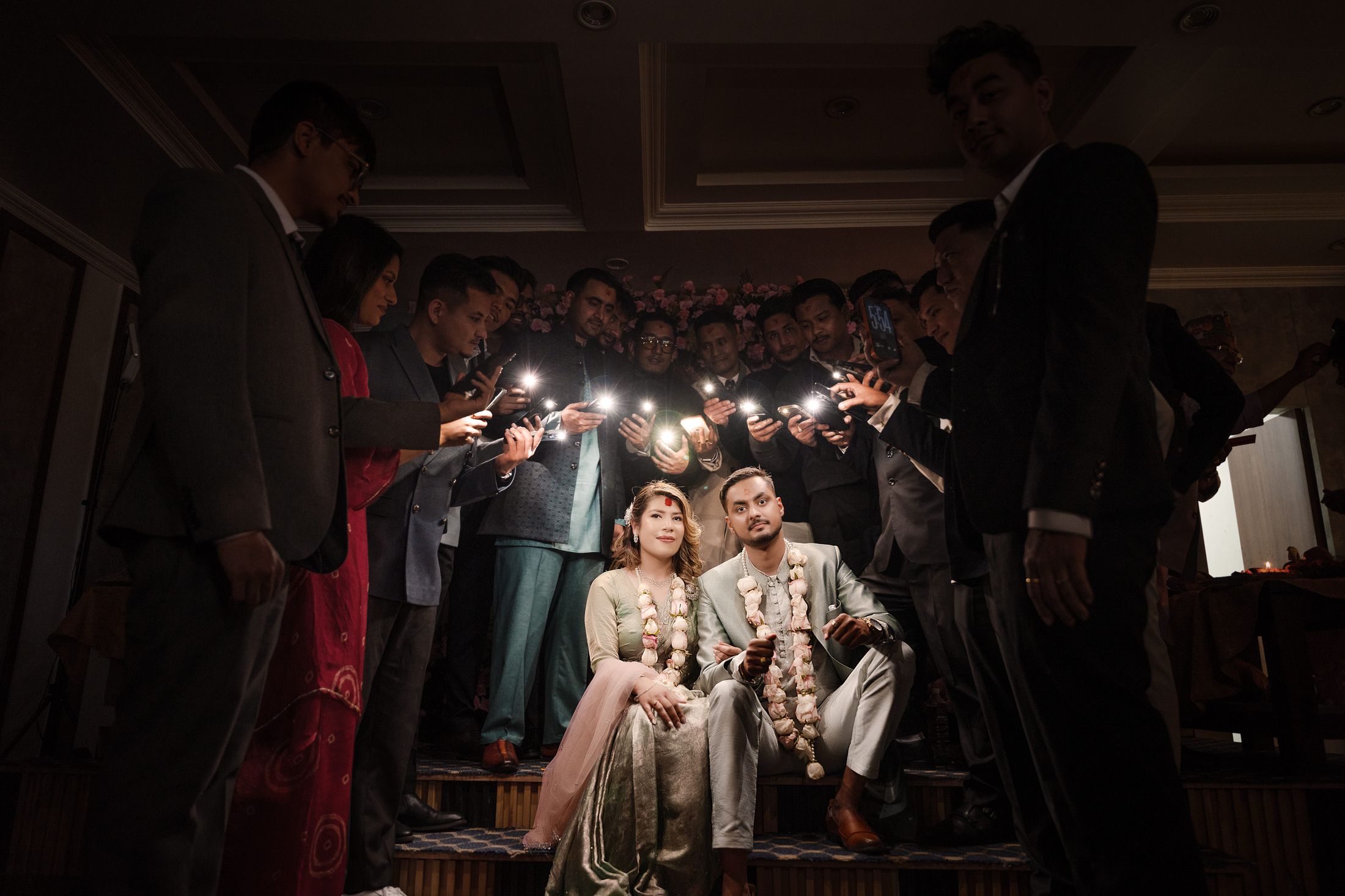 Photographers and guests with cameras and phones surrounding Suprabha and Nischal as they sit together wearing floral garlands