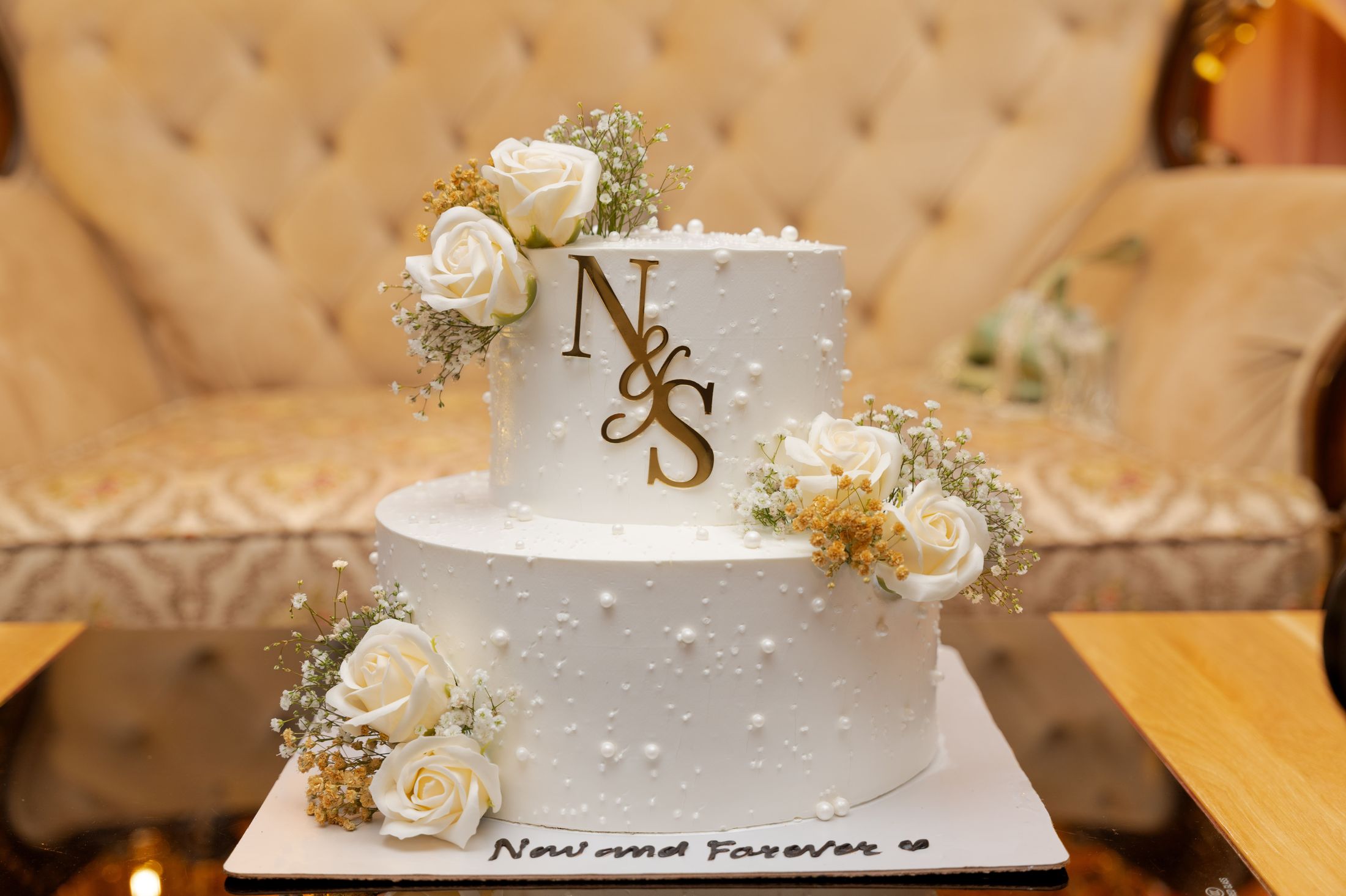 White two-tier wedding cake decorated with roses and a gold N&S monogram, inscribed Now and Forever