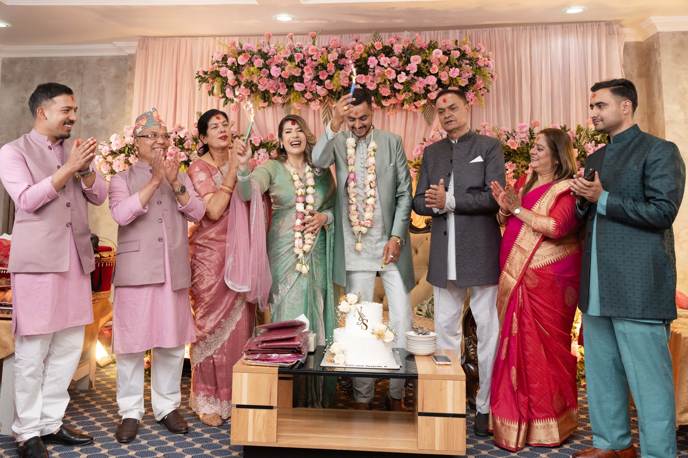 Family and friends clapping and celebrating around Suprabha and Nischal under a floral arch with the cake table in front