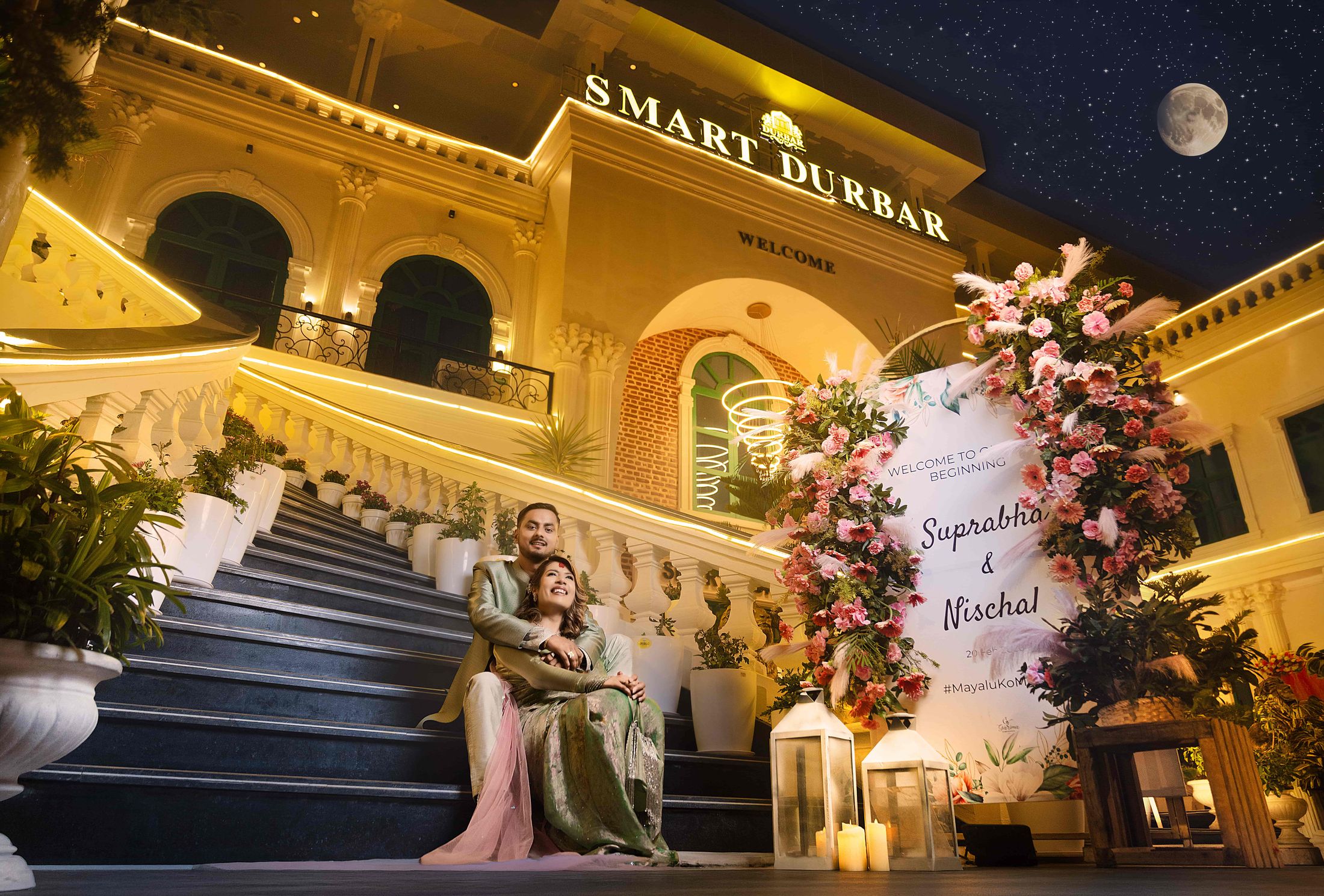 Suprabha and Nischal seated on an illuminated blue staircase outside the Smart Durbar venue beside a floral welcome sign with their names