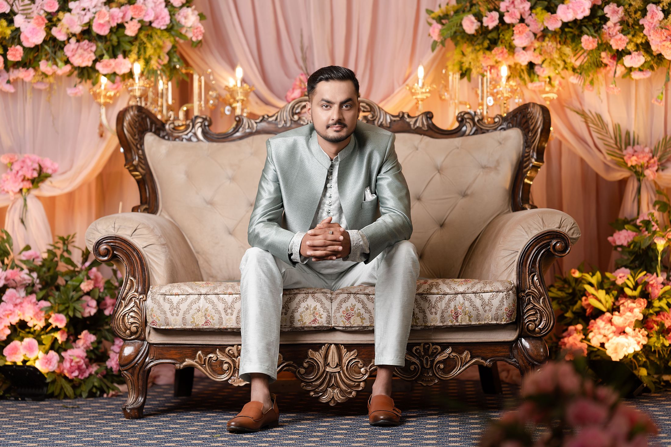 Nischal seated on an ornate tufted chair in a candlelit pink floral backdrop, hands clasped
