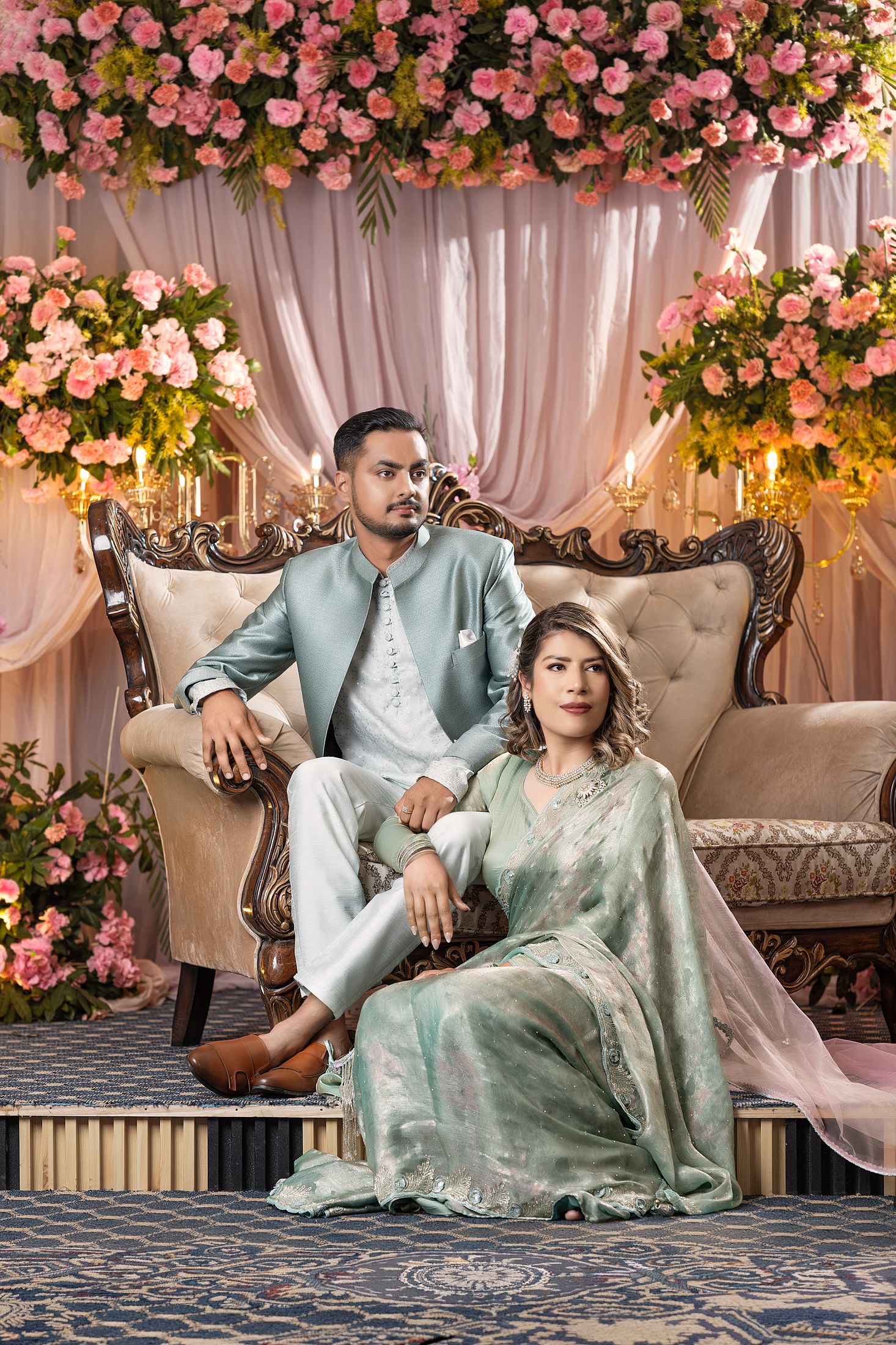 Suprabha and Nischal seated together on an ornate tufted chair, both dressed in matching sage green traditional outfits