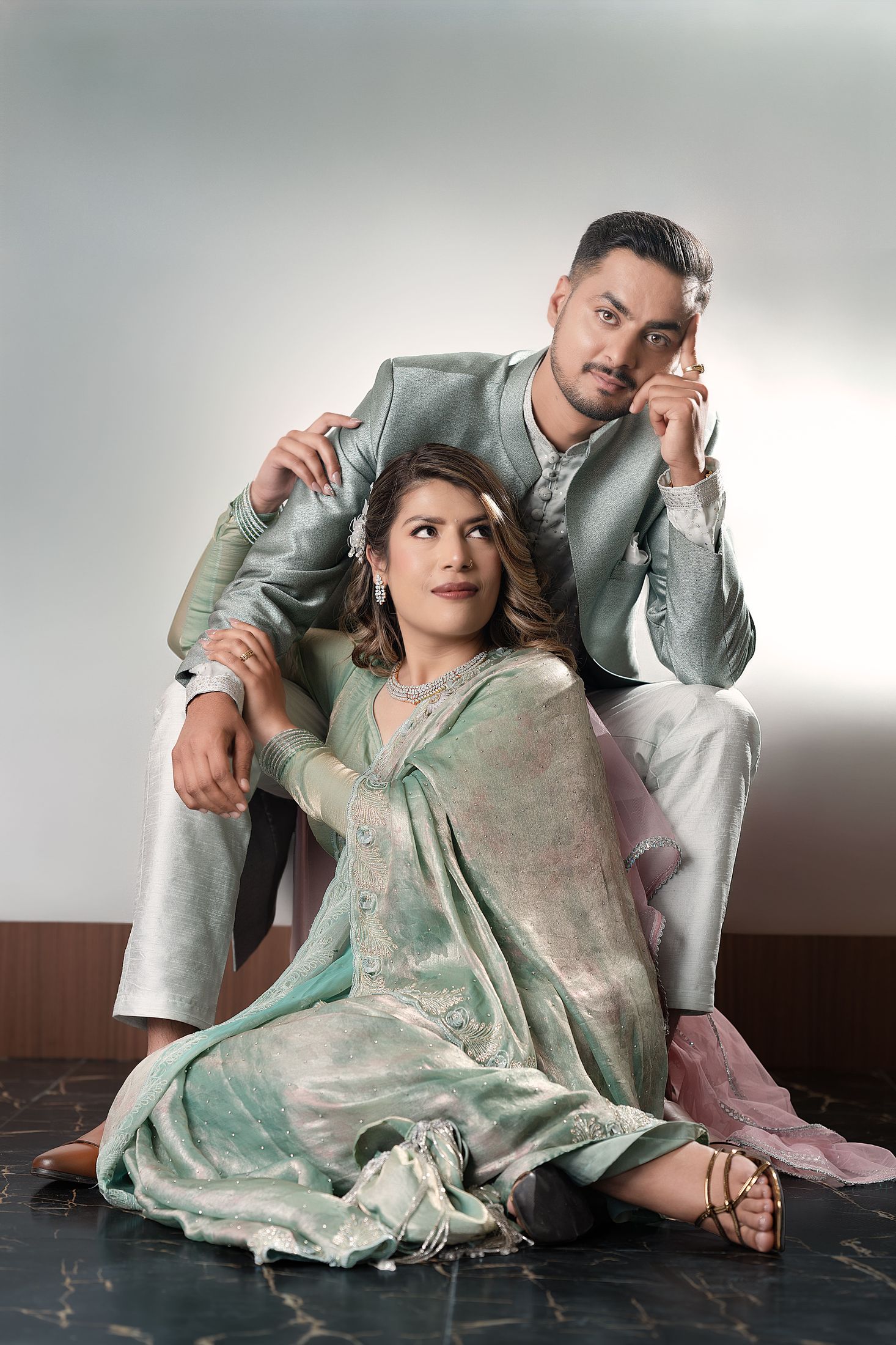 Suprabha seated on the floor in a flowing sage outfit with Nischal standing behind her, hand on her shoulder, against a grey studio background