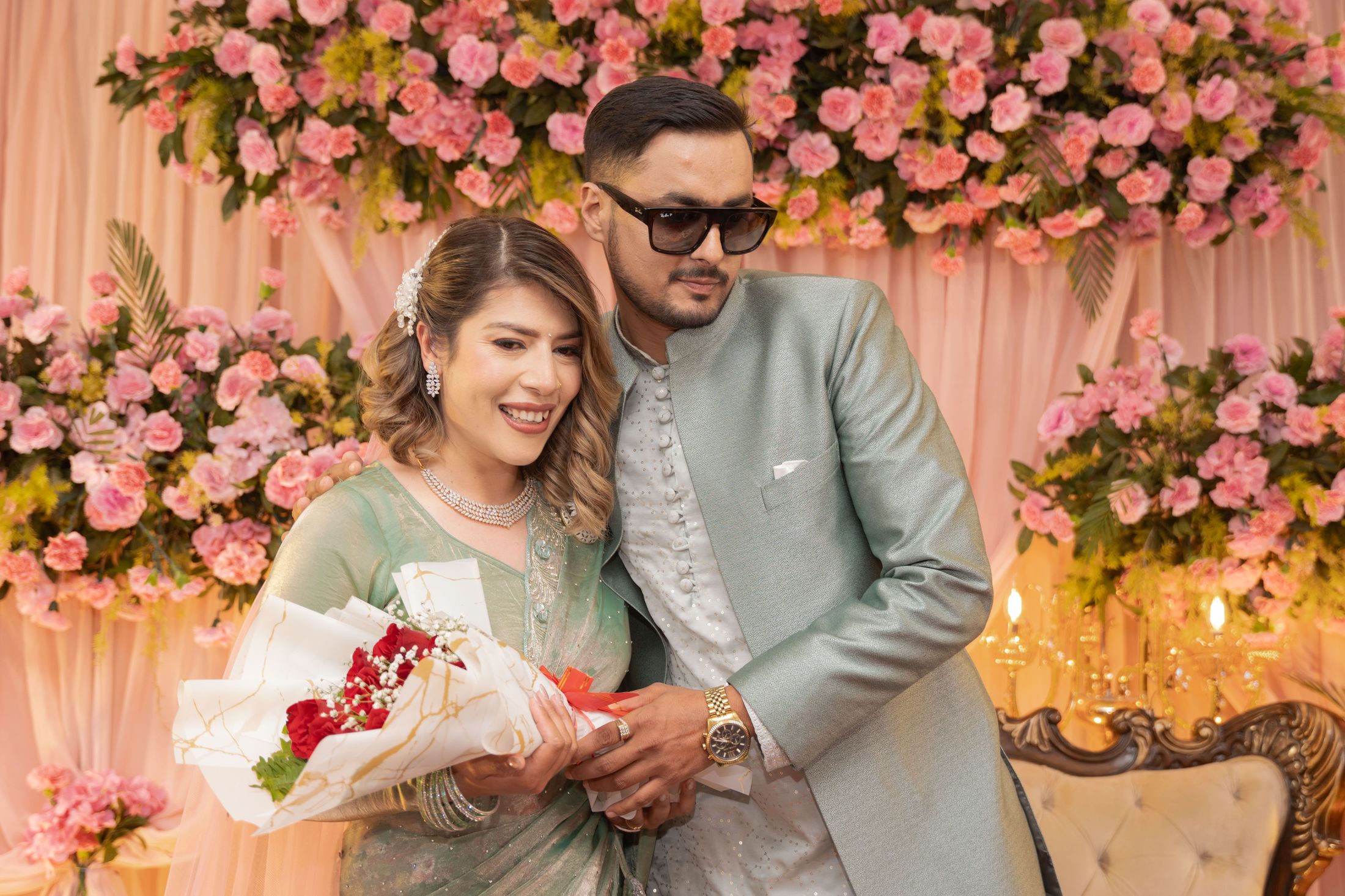 Suprabha and Nischal embracing in front of a pink rose floral backdrop, she holding a red bouquet and he wearing sunglasses