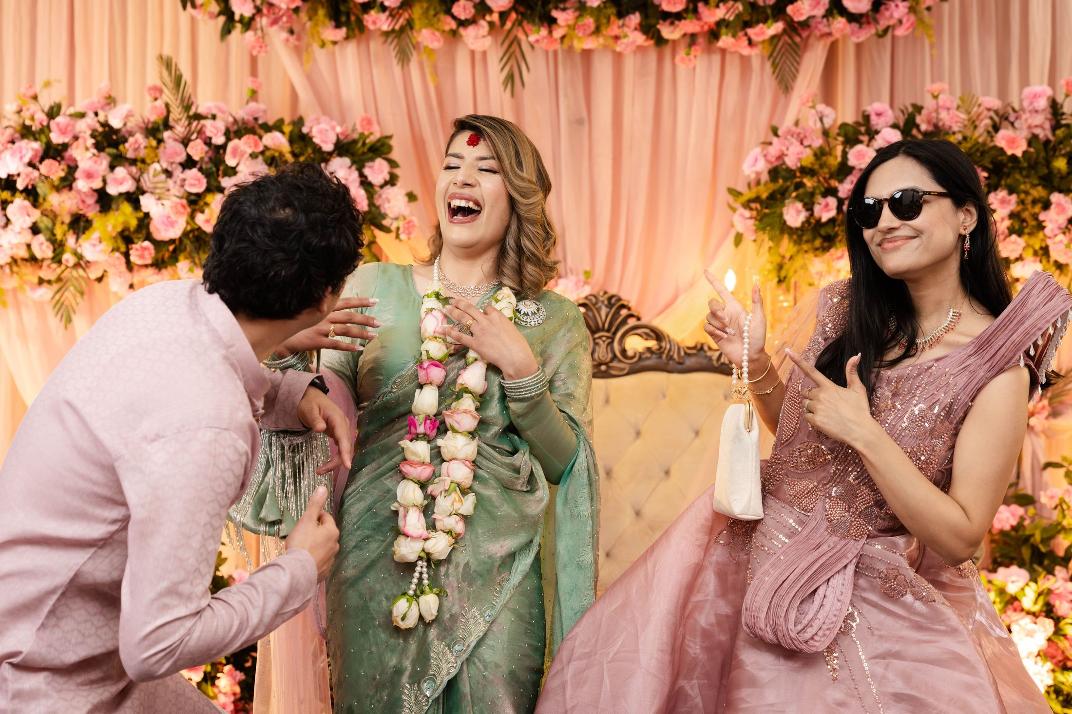 Suprabha laughing as Nischal kneels to adjust her floral garland while a friend in sunglasses looks on