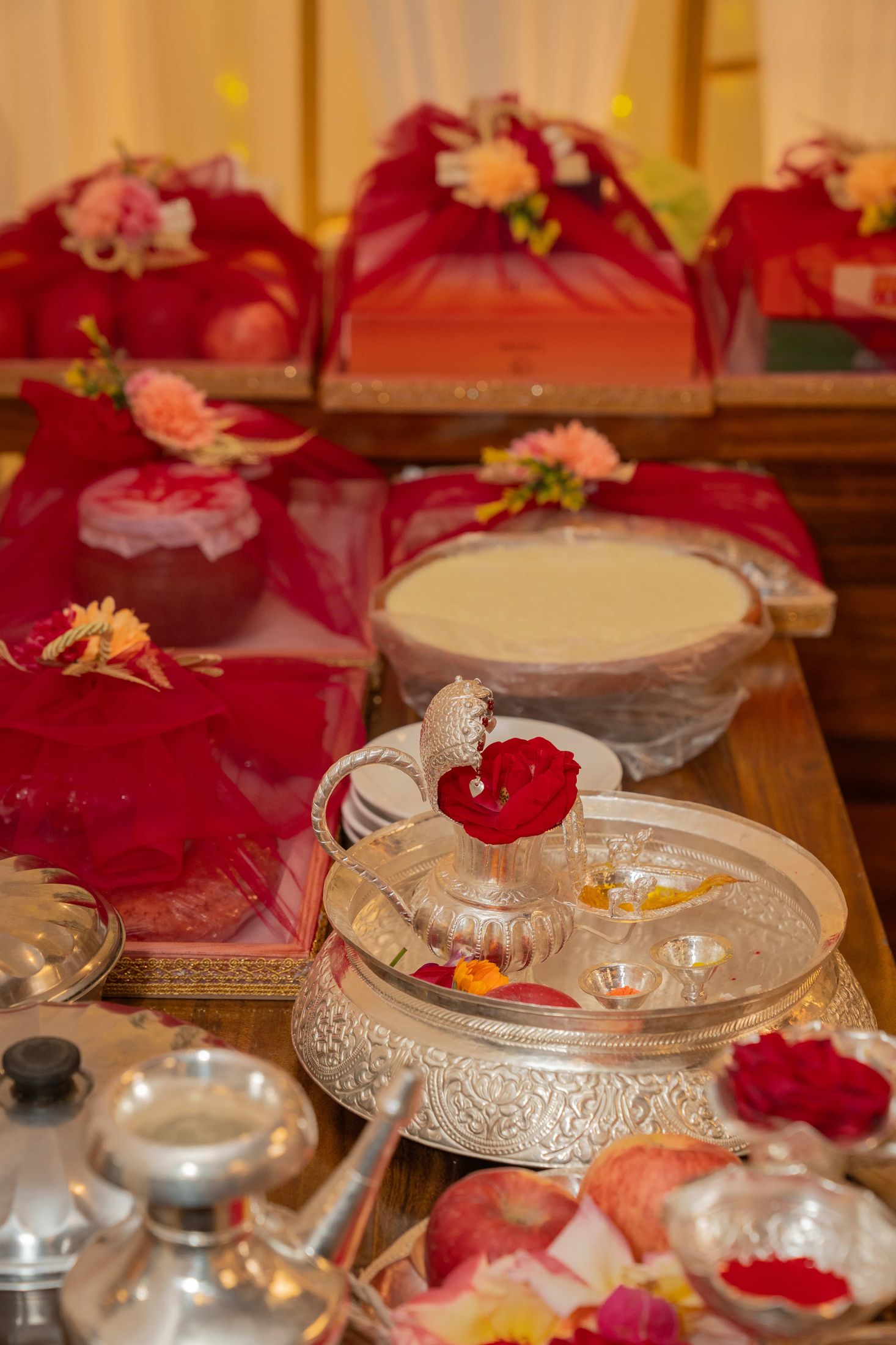 Red ceremonial trays with wrapped gifts and silver vessels holding tika and sindoor for the engagement ritual