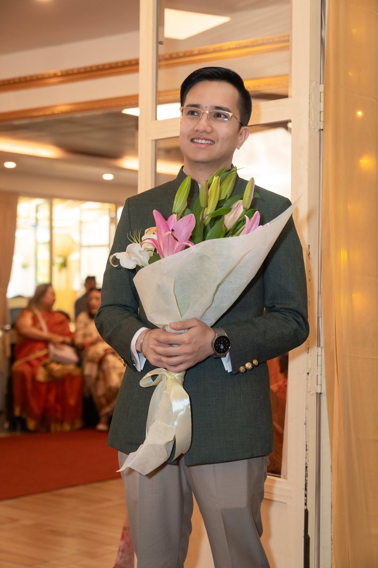 Samagya standing and holding a white-wrapped floral bouquet as he walks into the venue past seated guests