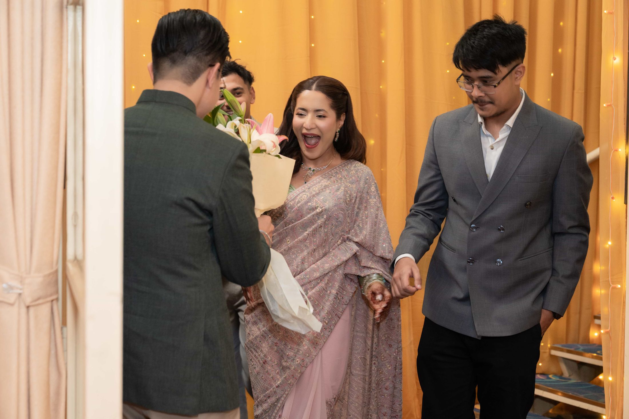 Samagya arriving with a bouquet as a woman in a pink saree reacts with joyful surprise, a man in a grey blazer standing beside her