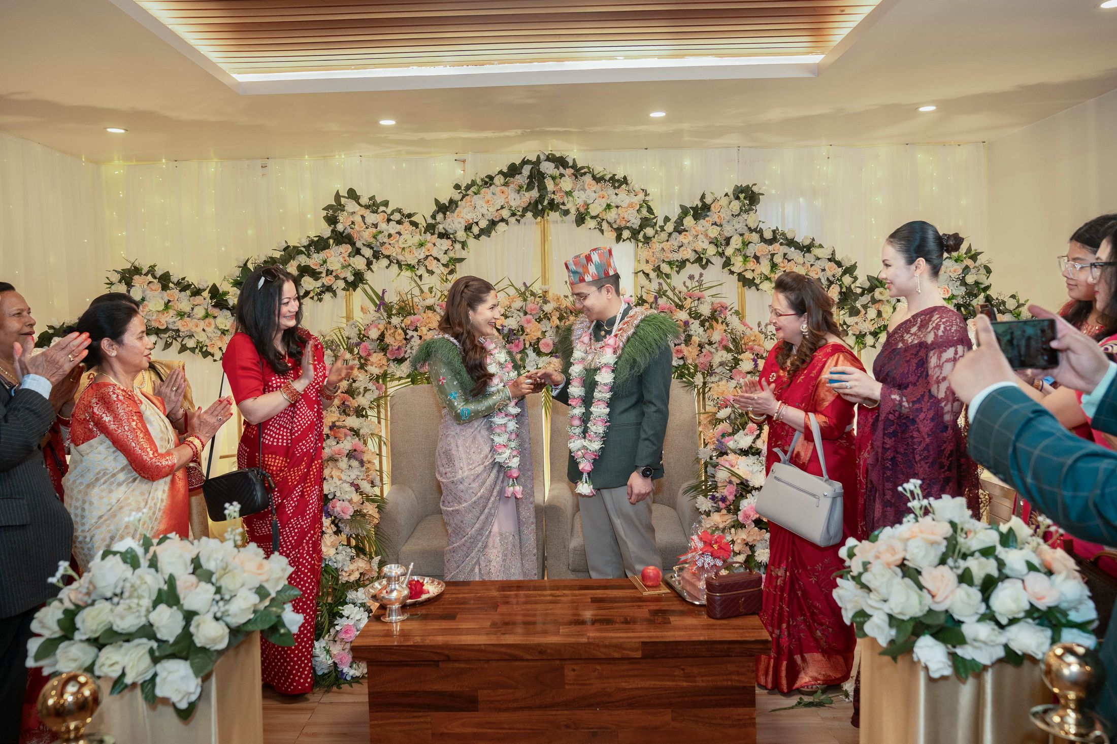 Shreya and Samagya exchanging garlands under a floral arch as guests clap and take photos around them