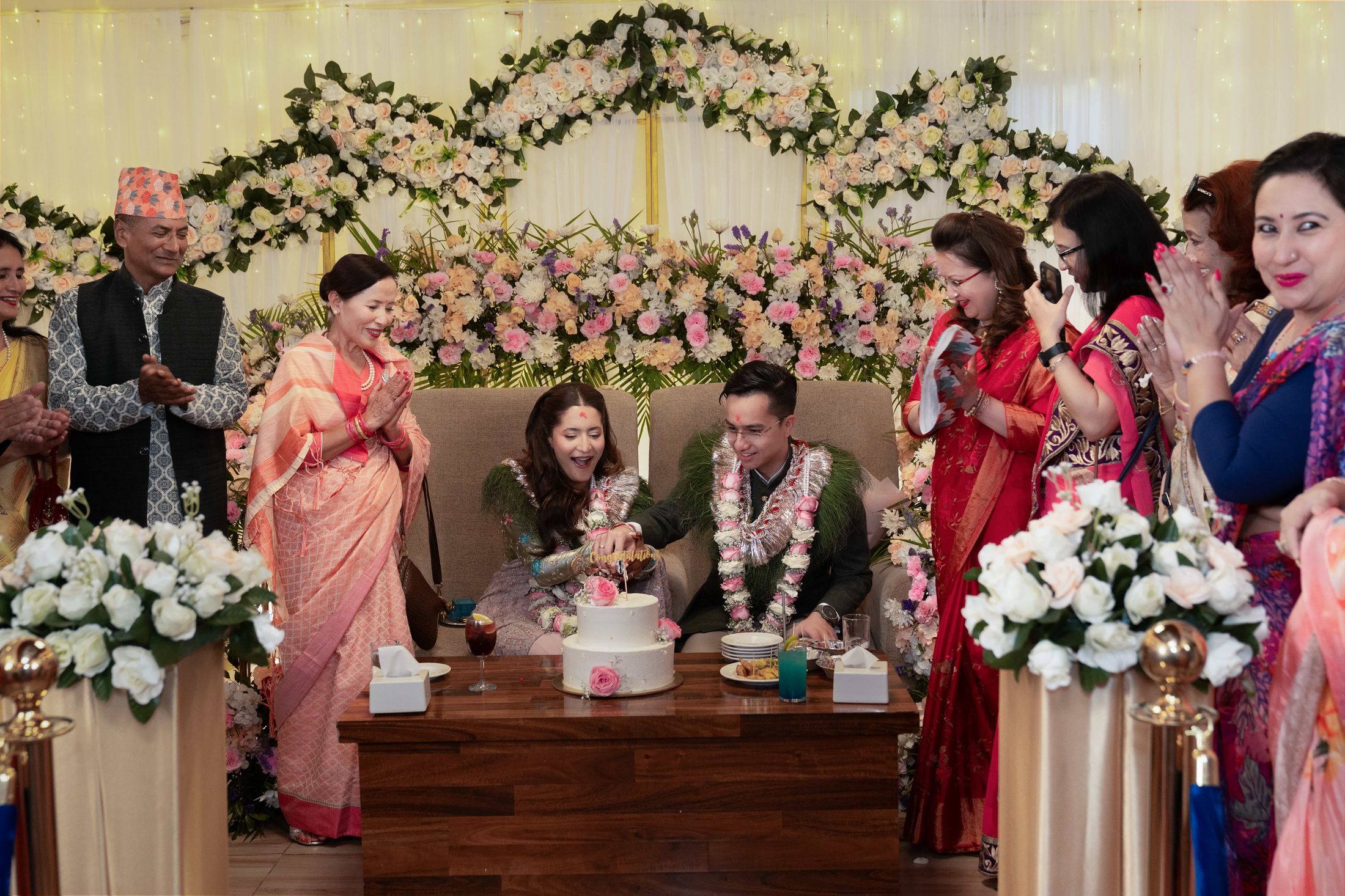 Family gathered around the cake-cutting table, groom's father in a vest and topi and women in sarees clapping and blessing the couple