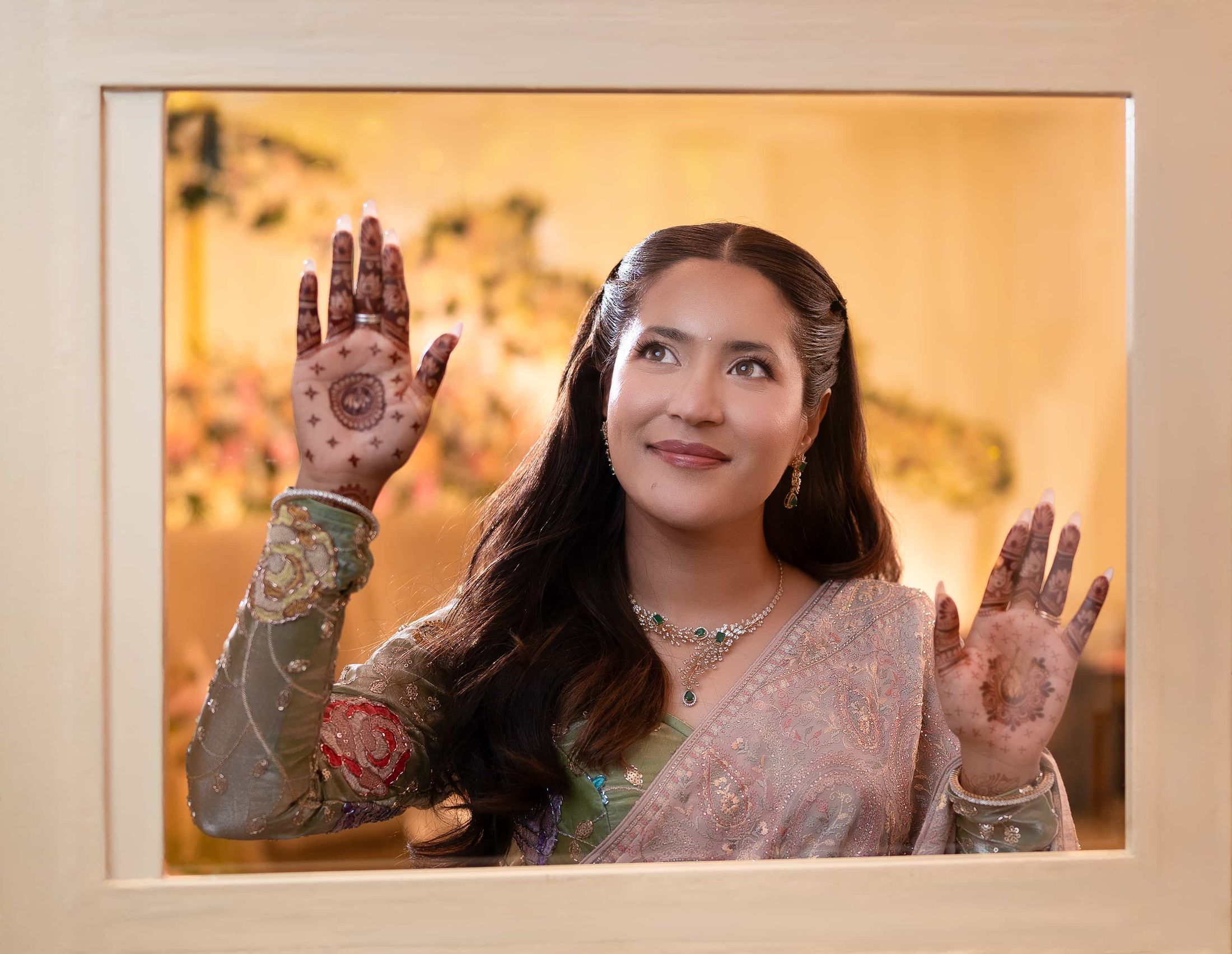 Shreya showing her mehendi-covered palms through a window frame, smiling in warm indoor lighting