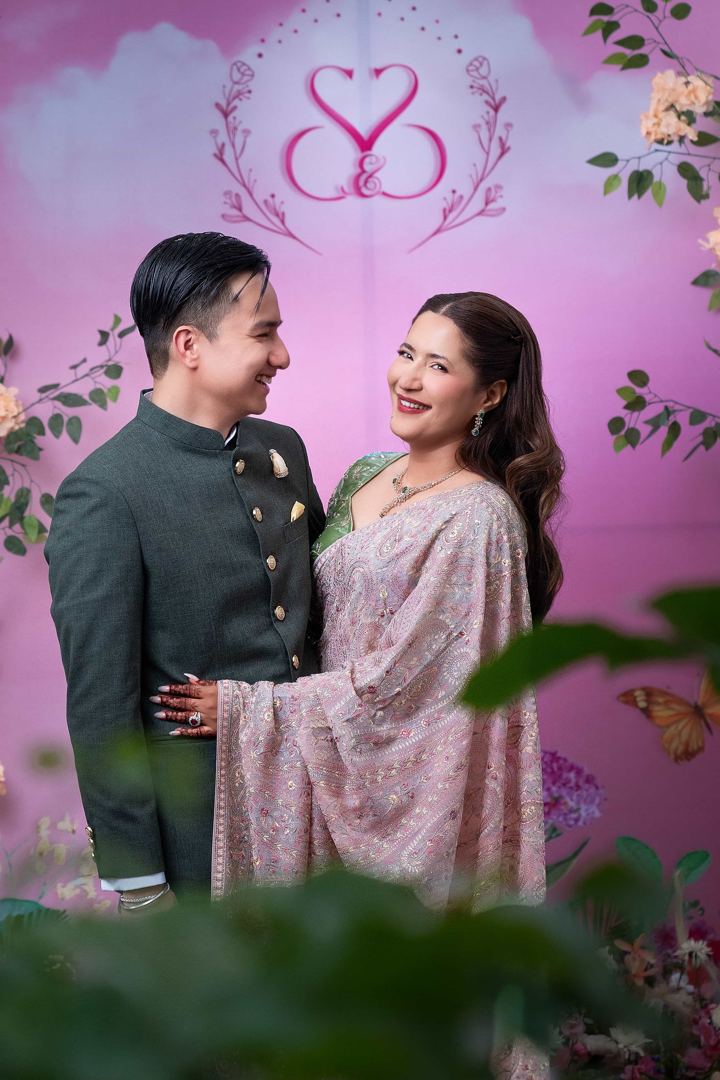 Shreya and Samagya embracing in front of a pink backdrop with their S&S monogram, greenery in the foreground